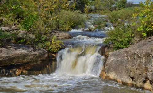 A serene waterfall cascades over rocks, surrounded by lush greenery and trees under a clear blue sky.