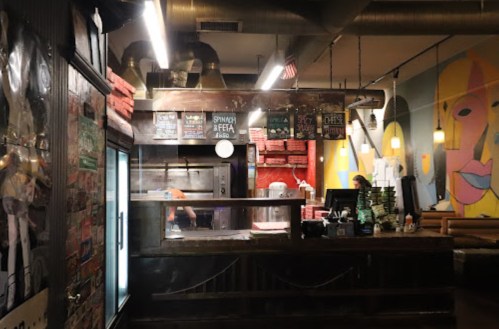 Interior of a cozy restaurant with a wooden counter, pizza boxes stacked, and colorful wall art.