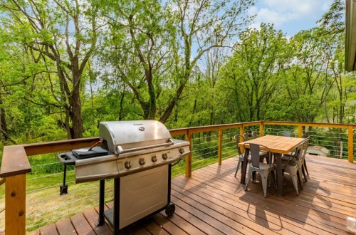 A spacious wooden deck with a grill and dining table, surrounded by lush green trees and a clear blue sky.