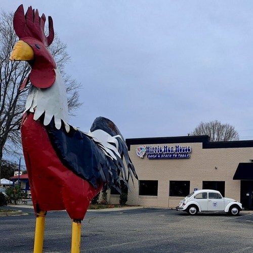 A large metal rooster sculpture stands in front of a restaurant with a vintage car parked nearby.