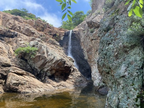 A serene waterfall cascading over rocky cliffs into a calm pool, surrounded by lush greenery and blue skies.