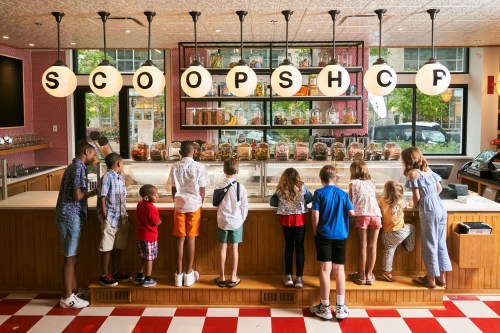 A group of children stands at a colorful ice cream shop counter, eagerly looking at the treats on display.