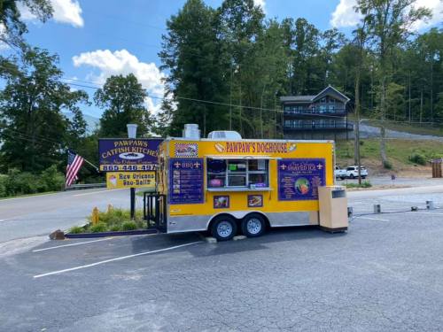 A colorful food truck named "Pawpaw's Doghouse" parked near a sign for "Pawpaw's Catfish Kitchen" in a scenic area.