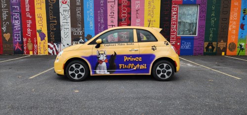 A yellow car with a colorful mural featuring a cat named "Prince Fluffytail" parked in front of a vibrant wall.