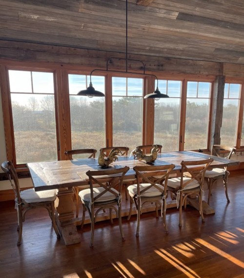 A rustic dining room with a large wooden table and chairs, featuring large windows that overlook a scenic view.