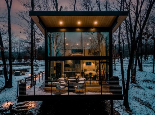 Modern glass house surrounded by trees, featuring a deck with seating, set in a snowy landscape at dusk.