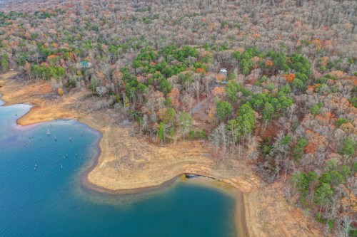 Aerial view of a forested area by a lake, showcasing autumn foliage and a sandy shoreline.