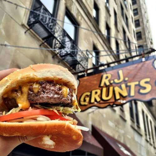 A hand holding a cheeseburger with lettuce and tomato in front of the R.J. Grunts restaurant sign.