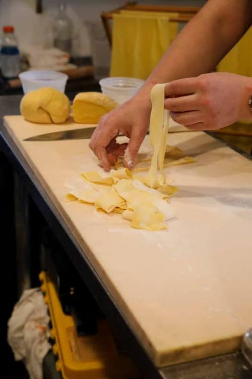 A person’s hands cutting fresh pasta on a wooden countertop, with dough and flour visible in the background.