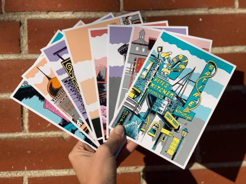 A hand holding a fan of colorful postcards featuring various illustrated cityscapes against a brick background.