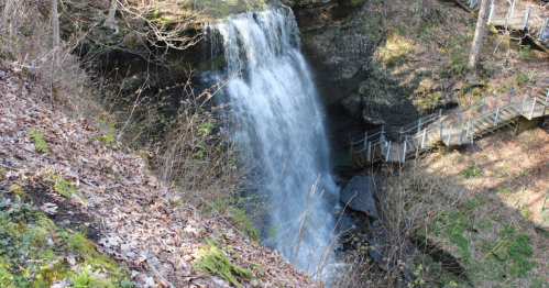 A waterfall cascading down rocky terrain, surrounded by greenery and a wooden pathway nearby.
