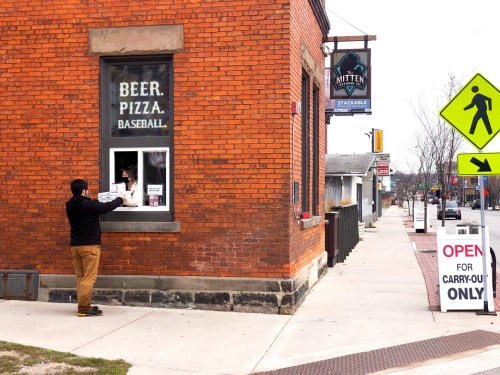 A person at a takeout window orders from a restaurant with a sign that reads "Beer. Pizza. Baseball." on a city street.