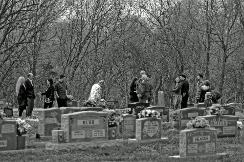 A group of people gathered in a cemetery, surrounded by gravestones and bare trees, in a somber atmosphere.