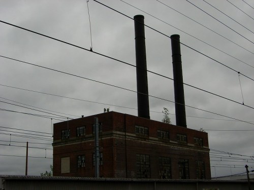 A brick industrial building with two tall smokestacks against a cloudy sky, surrounded by power lines.