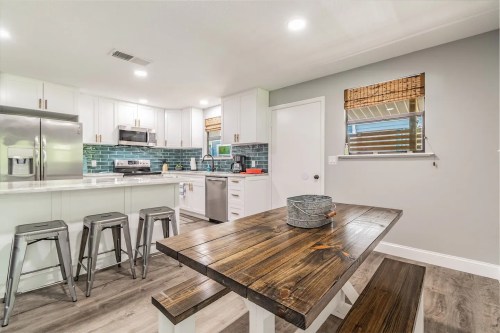 Modern kitchen with stainless steel appliances, a wooden dining table, and blue tile backsplash. Bright and inviting space.