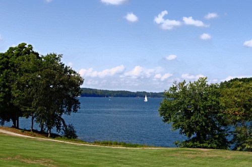 A serene lake view with a sailboat, surrounded by green trees and a clear blue sky with fluffy clouds.