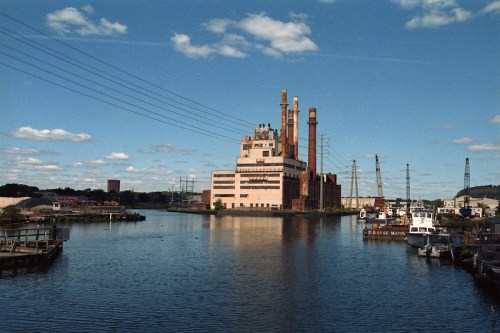 Industrial building with smokestacks by a calm river, surrounded by boats and cranes under a clear blue sky.