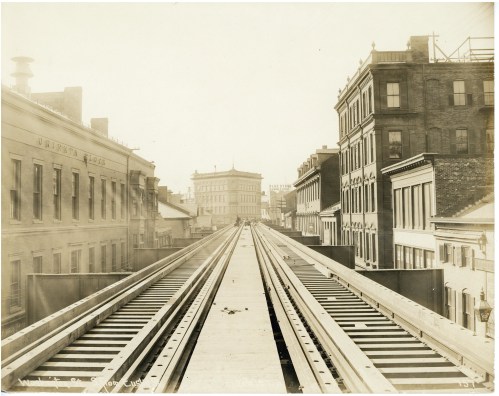Black and white photo of elevated train tracks running through a city, flanked by historic buildings.