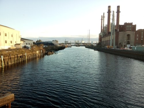 A calm waterway with industrial buildings and power plant smokestacks in the background, under a clear sky.