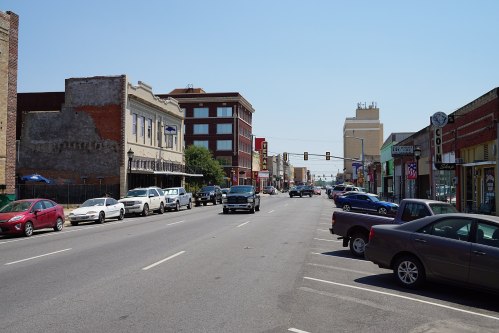 A view of a downtown street lined with buildings and parked cars under a clear blue sky. Traffic lights visible ahead.