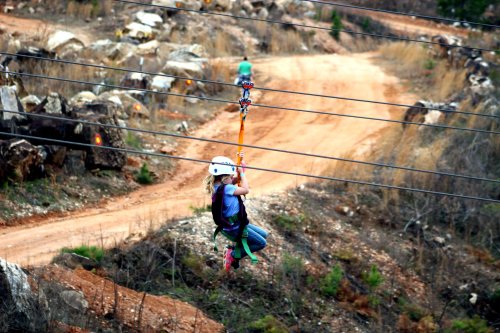 A child in a helmet zip-lining over a dirt path surrounded by rocky terrain and sparse vegetation.