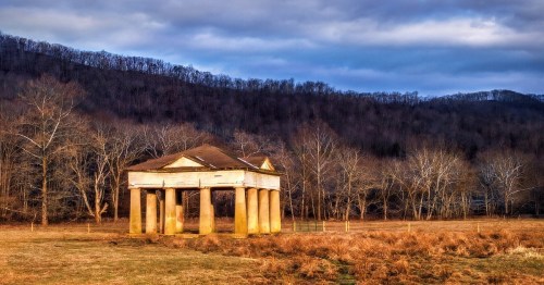 An abandoned structure with columns stands in a grassy field, surrounded by bare trees and rolling hills under a cloudy sky.
