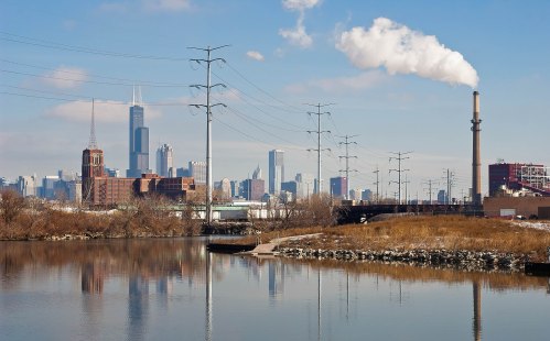 City skyline with skyscrapers, power lines, and a smokestack reflected in a calm river under a clear blue sky.