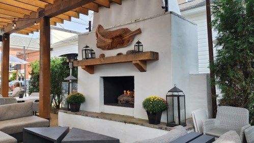 Cozy outdoor seating area with a fireplace, wooden beams, and decorative lanterns, surrounded by greenery and flowers.