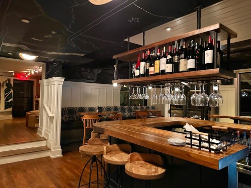 A cozy bar area with wooden stools, a wine rack overhead, and a stylish black ceiling with a map design.