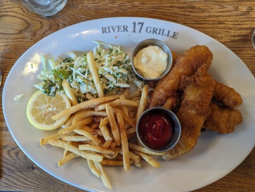 A plate of fried fish, fries, coleslaw, lemon, and dipping sauces at River 17 Grille.