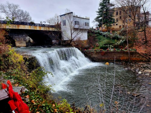 A waterfall cascades under a bridge, surrounded by autumn foliage and a quaint building in the background.