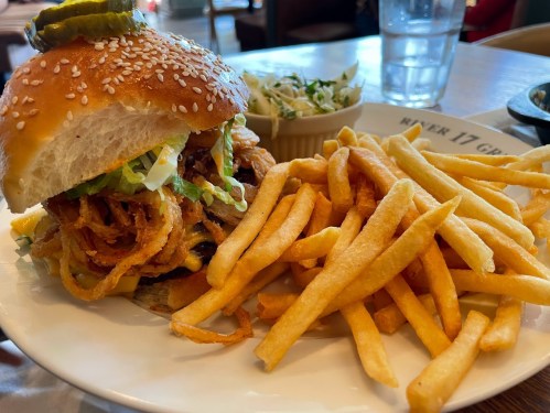A sesame seed burger topped with lettuce, pickles, and crispy onions, served with fries and a side of coleslaw.