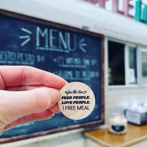 A hand holds a wooden token that reads "For the love, feed people. Love people. 1 free meal." in front of a menu board.