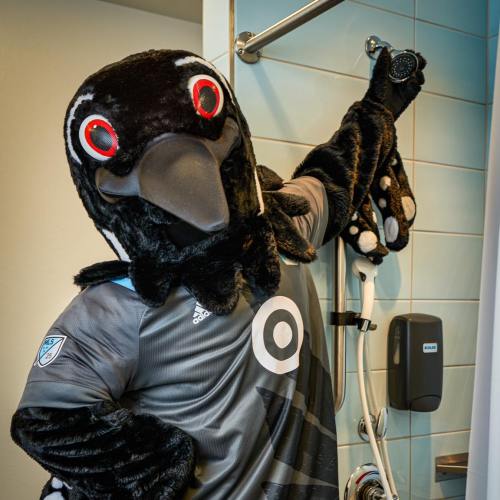 A mascot in a black bird costume poses playfully in a bathroom, holding a showerhead.