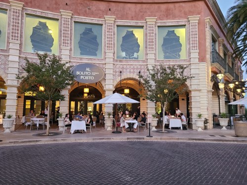 Outdoor dining scene at a restaurant with patrons seated at tables under umbrellas, surrounded by palm trees.