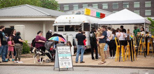 A crowd gathers outside a food truck, with people chatting, a dog on a leash, and colorful decorations overhead.
