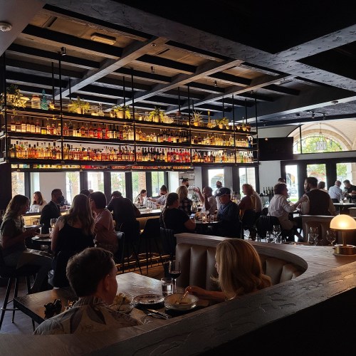 A bustling restaurant interior with patrons dining, a bar lined with bottles, and large windows letting in natural light.