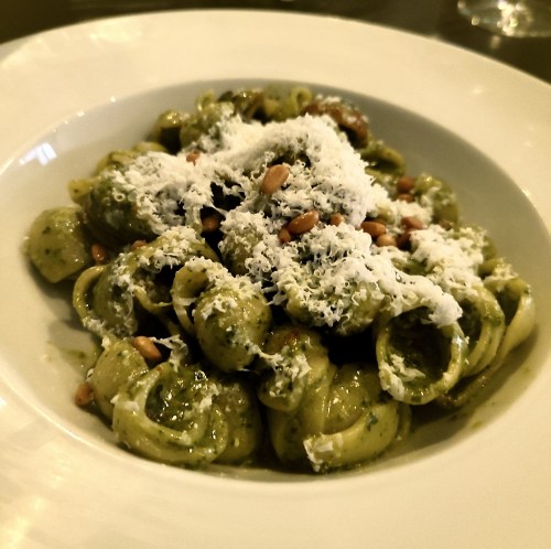 A plate of pasta with pesto, topped with grated cheese and pine nuts, served in a white bowl.