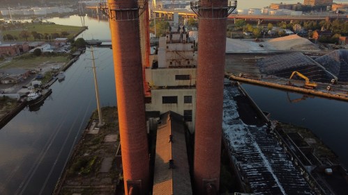 Aerial view of an industrial site with tall smokestacks, a canal, and construction materials in the background.