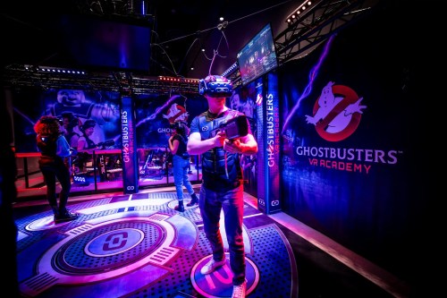A person wearing a VR headset stands in a colorful Ghostbusters VR Academy setup, surrounded by themed decorations.