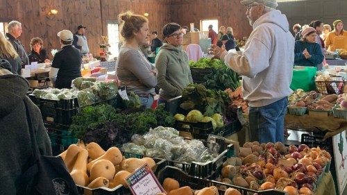 A bustling farmers market with vendors and customers discussing fresh produce and goods. Various vegetables are displayed.