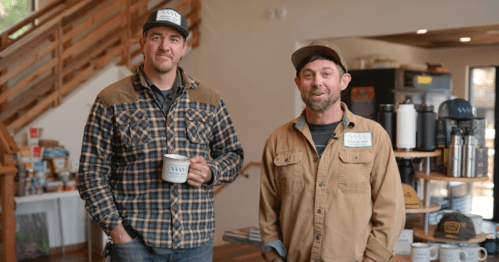 Two men stand in a cozy shop, smiling. One holds a coffee mug, both wear casual hats and shirts.