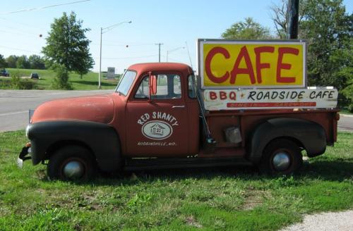 An old red pickup truck parked by the roadside with a sign reading "Cafe BBQ Roadside Cafe."