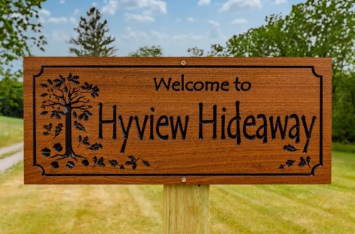Wooden sign reading "Welcome to Hyview Hideaway" surrounded by greenery and a clear blue sky.