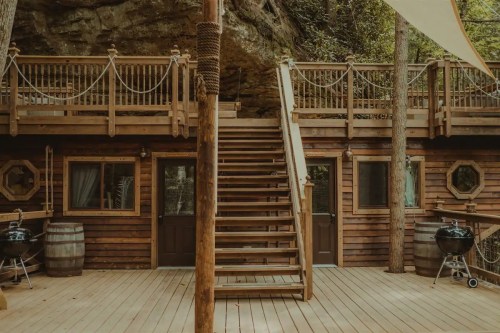 Wooden deck with stairs leading up to a rustic cabin, surrounded by trees and featuring outdoor grills.