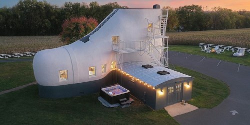 A whimsical shoe-shaped house with a staircase, surrounded by greenery and a hot tub, at sunset.