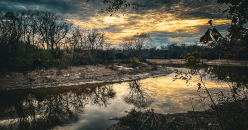 A serene river scene at sunset, with trees reflecting on the water and a dramatic sky filled with clouds.
