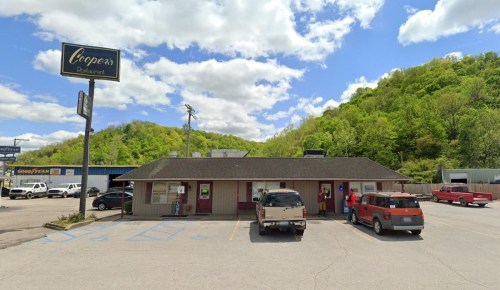 A restaurant building with a sign, surrounded by green hills and parked cars under a partly cloudy sky.