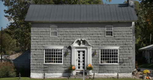 A charming gray house with a shingled exterior, decorative front door, and pumpkins on the porch.