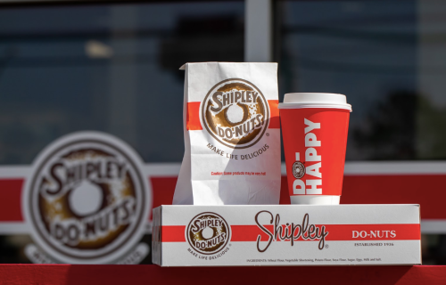 A Shipley Do-Nuts bag and coffee cup sit on a table, with the shop's logo in the background.